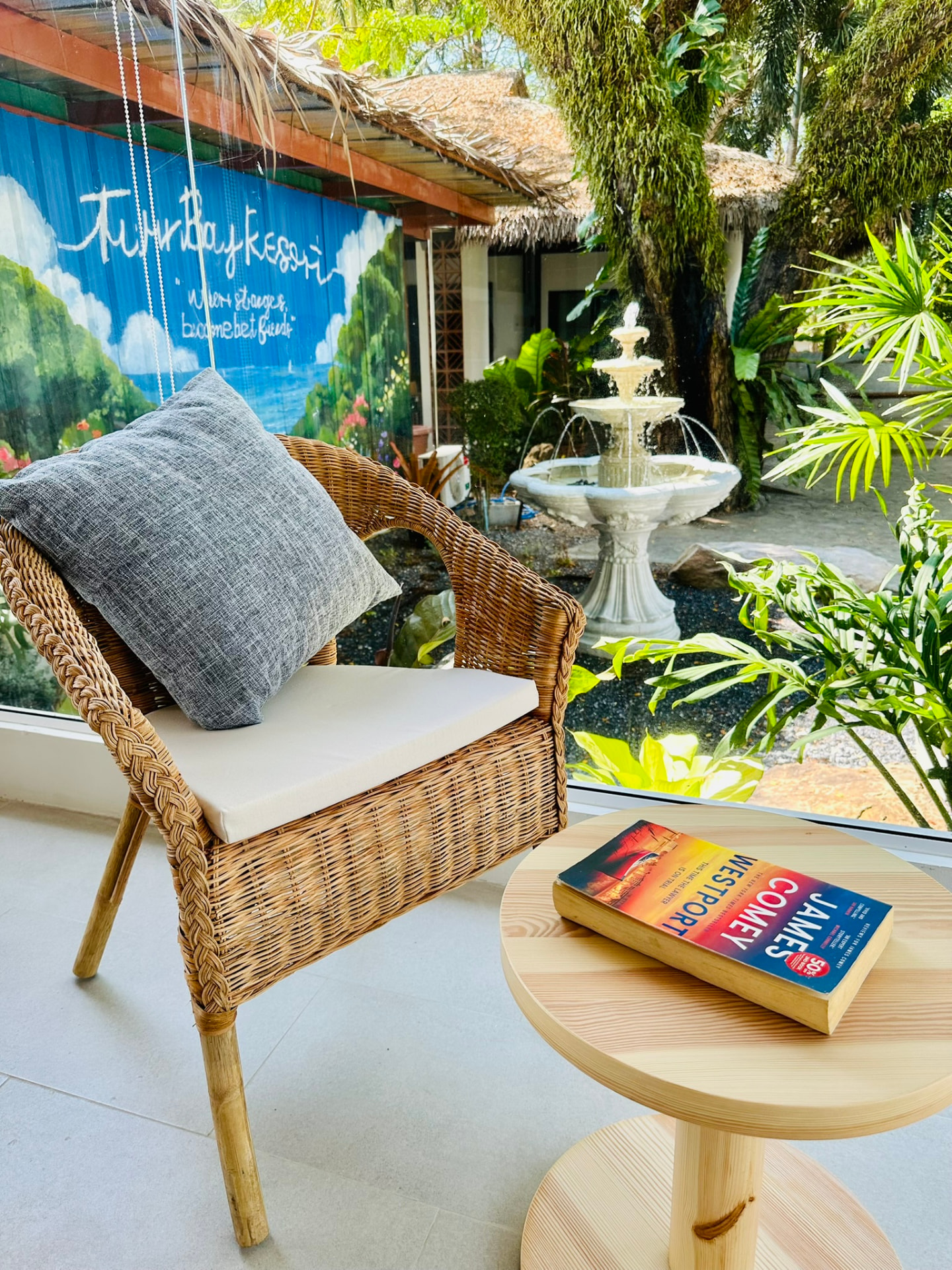 A rattan reading chair beside a book on a side table, with the garden fountain outside