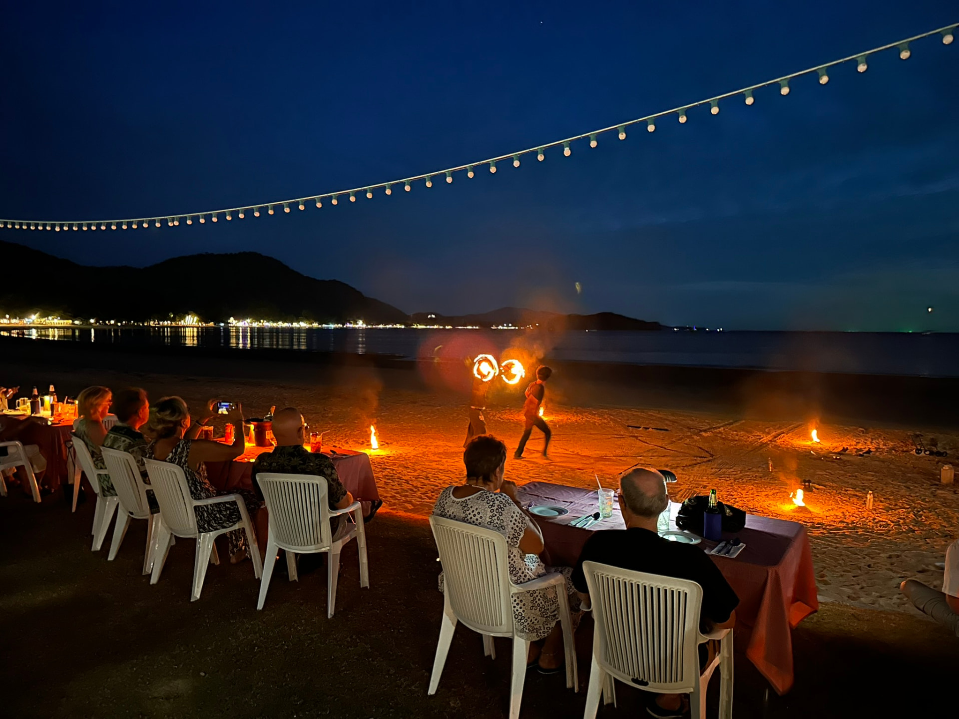 A fire dancer spinning burning poi on the sand at dusk with guests watching from their tables