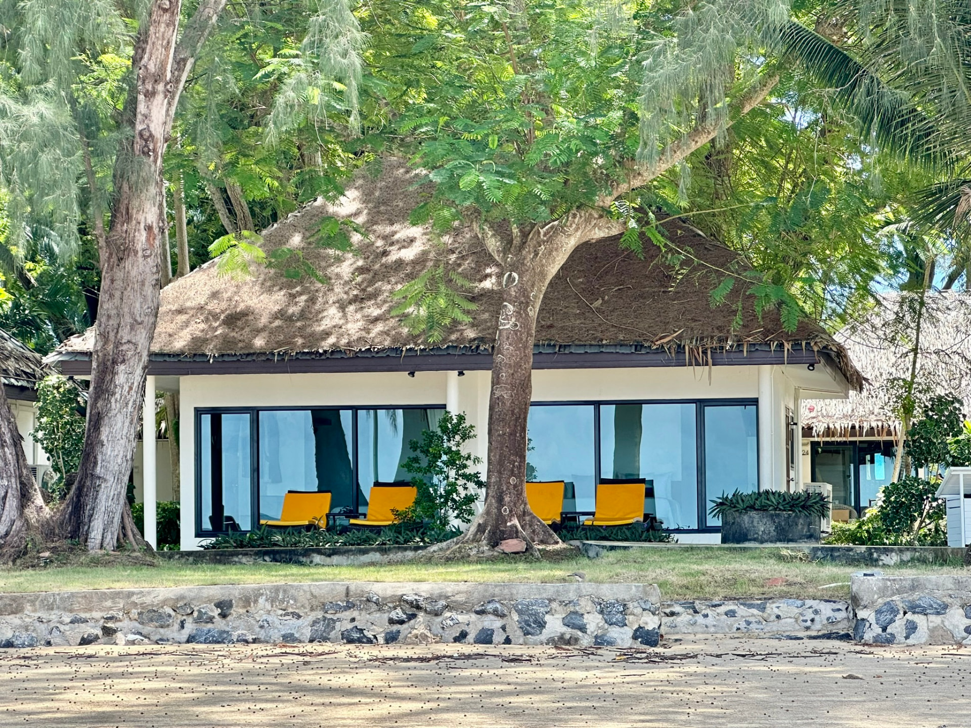 Beachfront bungalow seen from the sand, shaded by trees with yellow chairs on the veranda