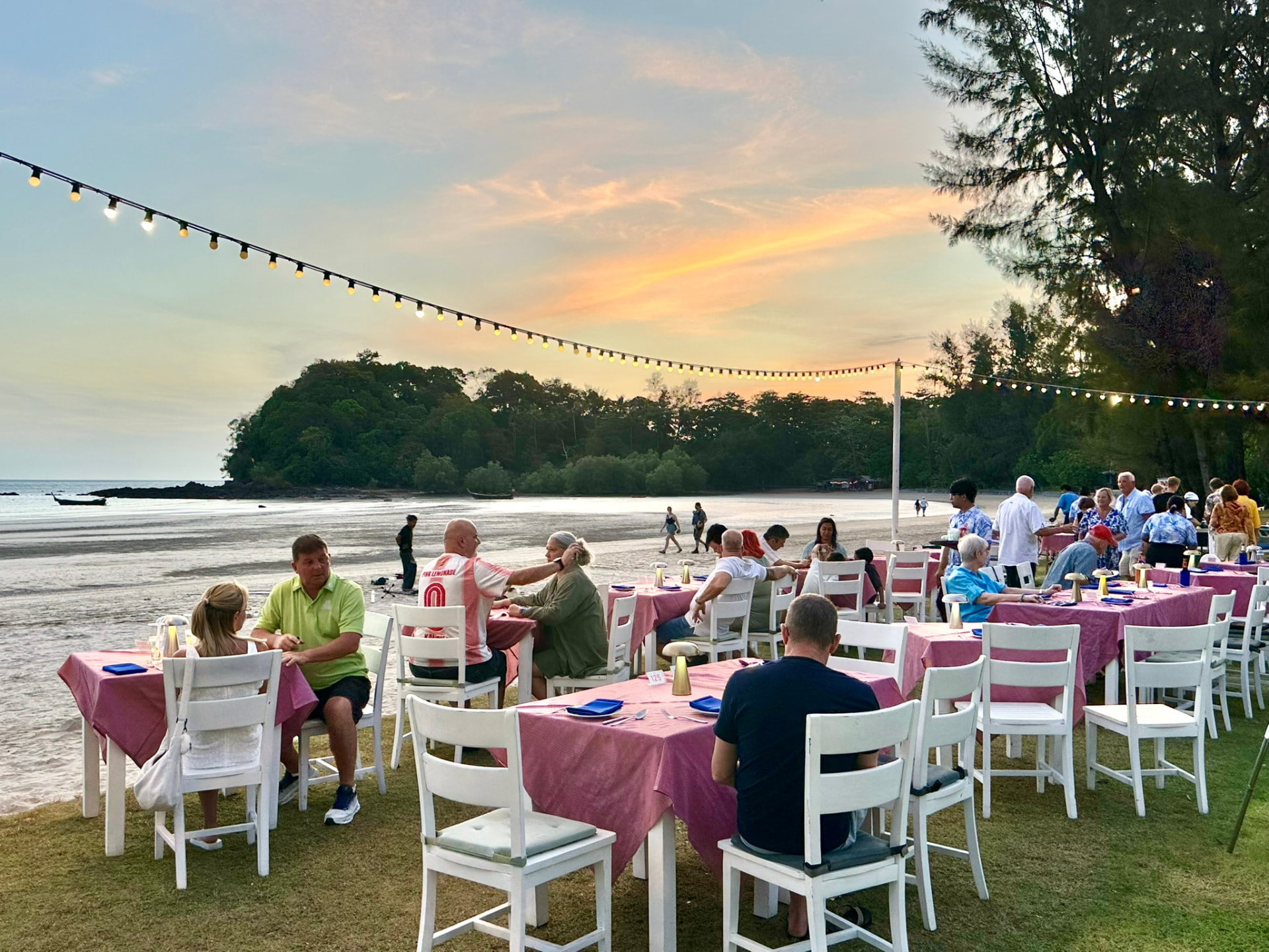 Long rows of tables under festoon lights on the beach at sunset before the BBQ dinner