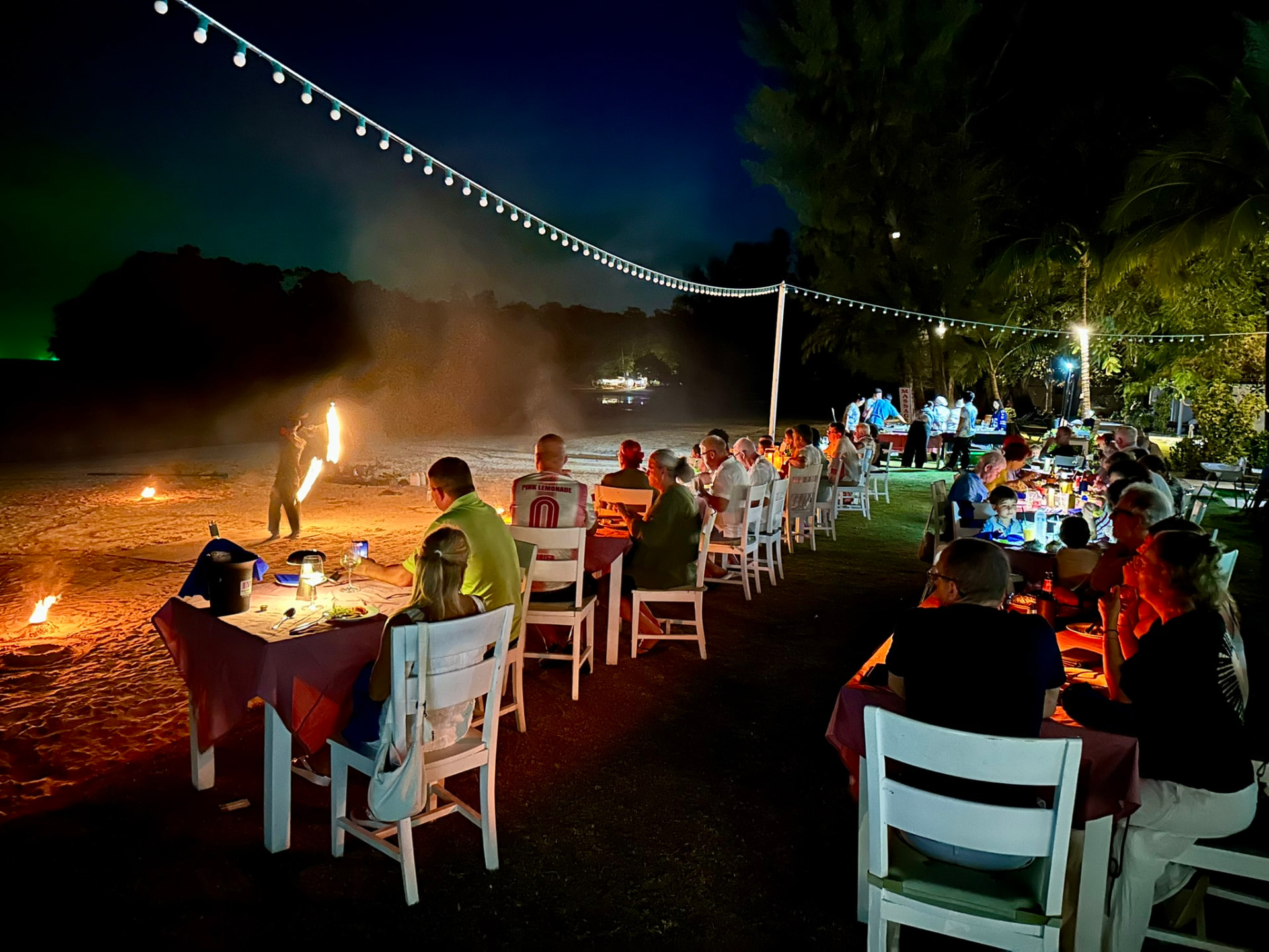 Guests dining at candlelit tables on the beach while a fire dancer performs beside the water