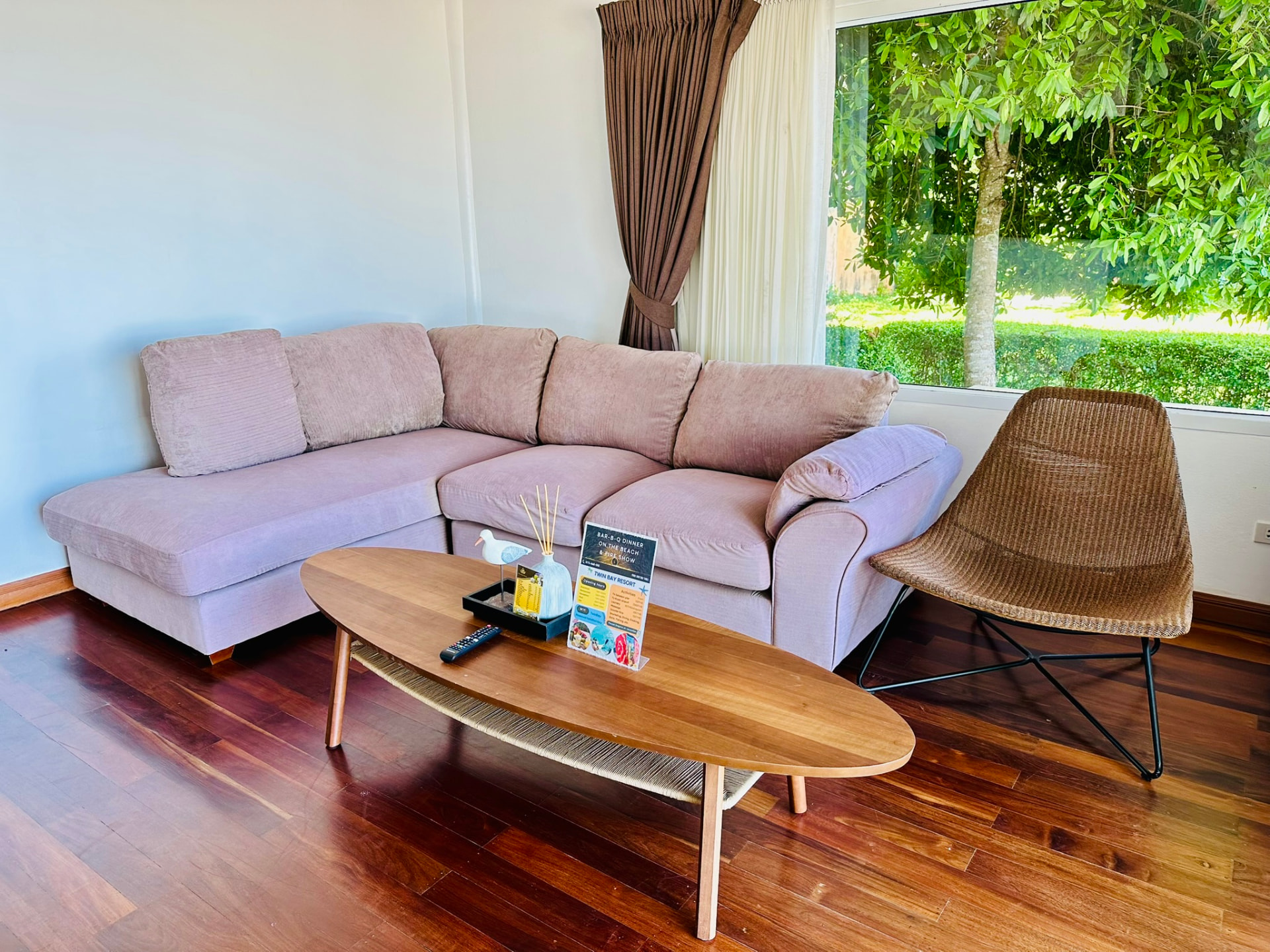 Corner sofa, oval coffee table and rattan chair beside a window full of greenery