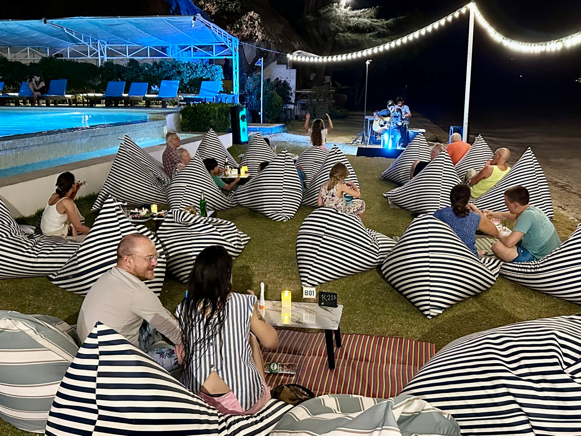 Guests on bean bags beside the lit pool watching a musician play under festoon lights