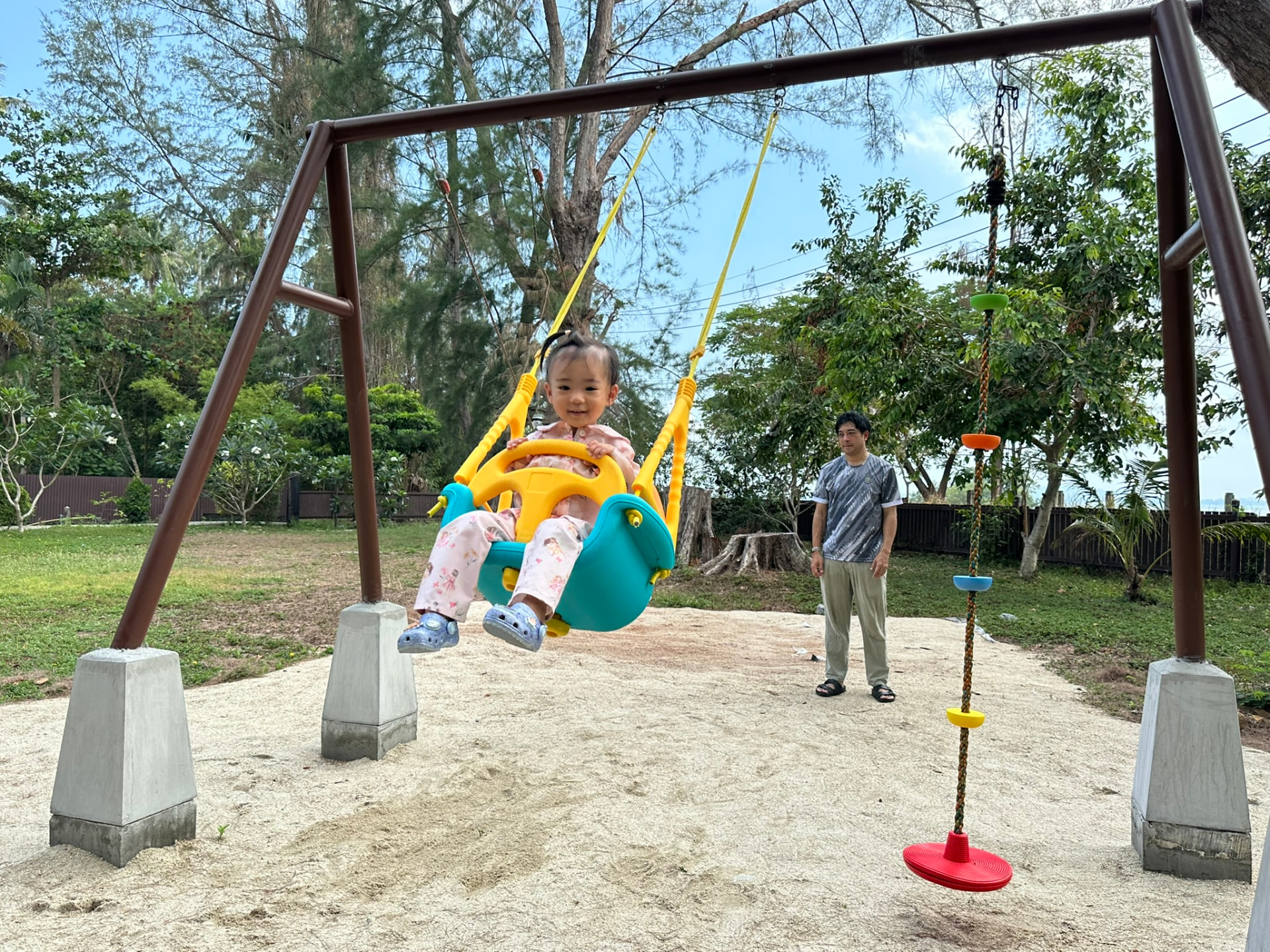A toddler laughing in the baby swing while a parent watches from the sand