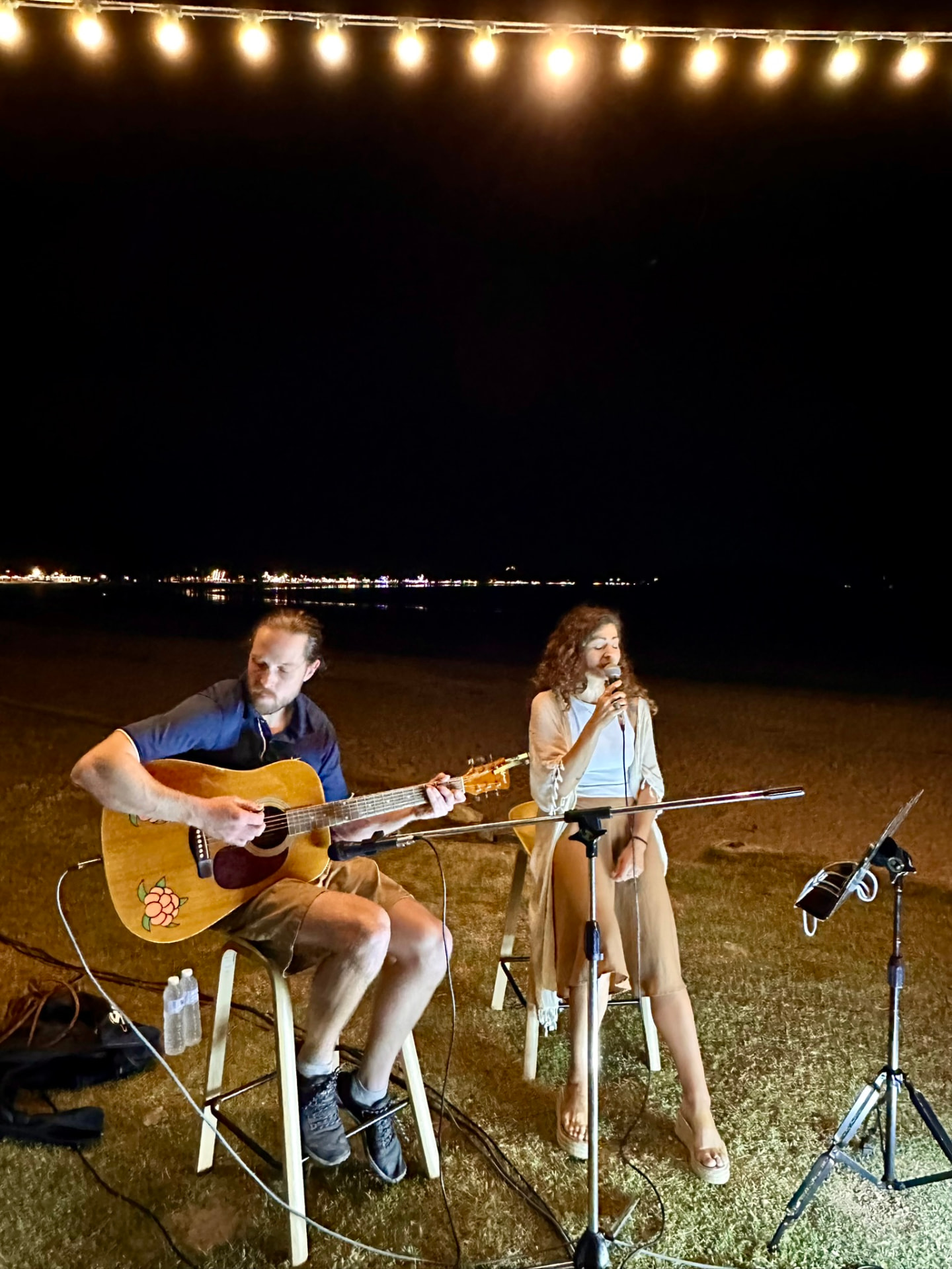 A guitarist and a singer performing on the beach under a string of warm lights