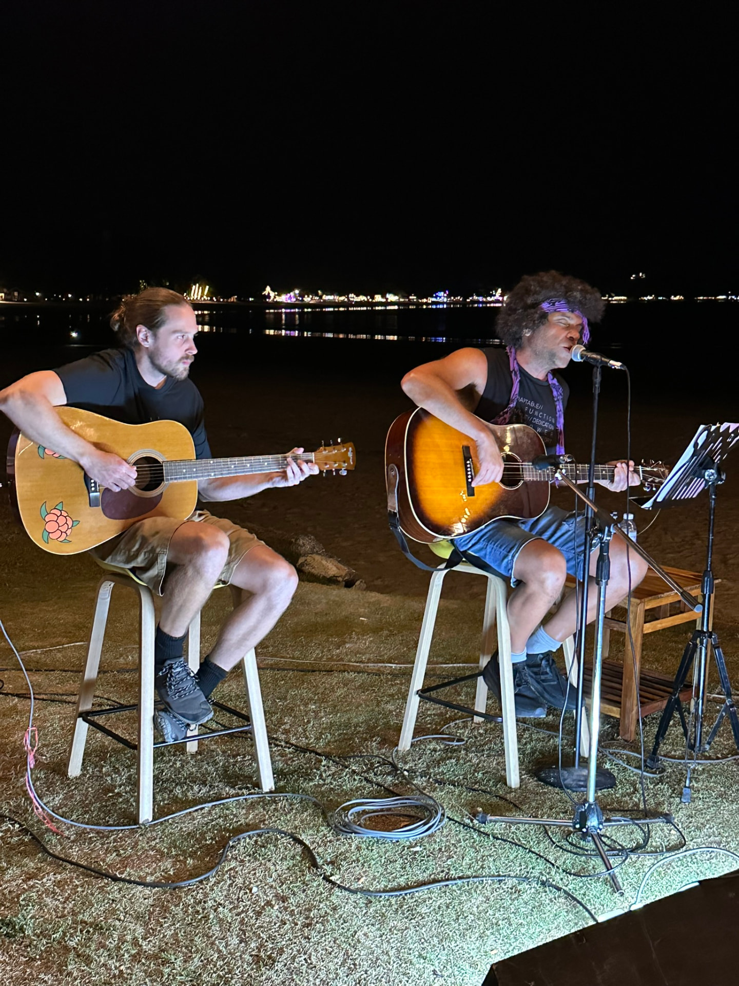 Two guitarists playing side by side on the beach at night with the bay lights behind them
