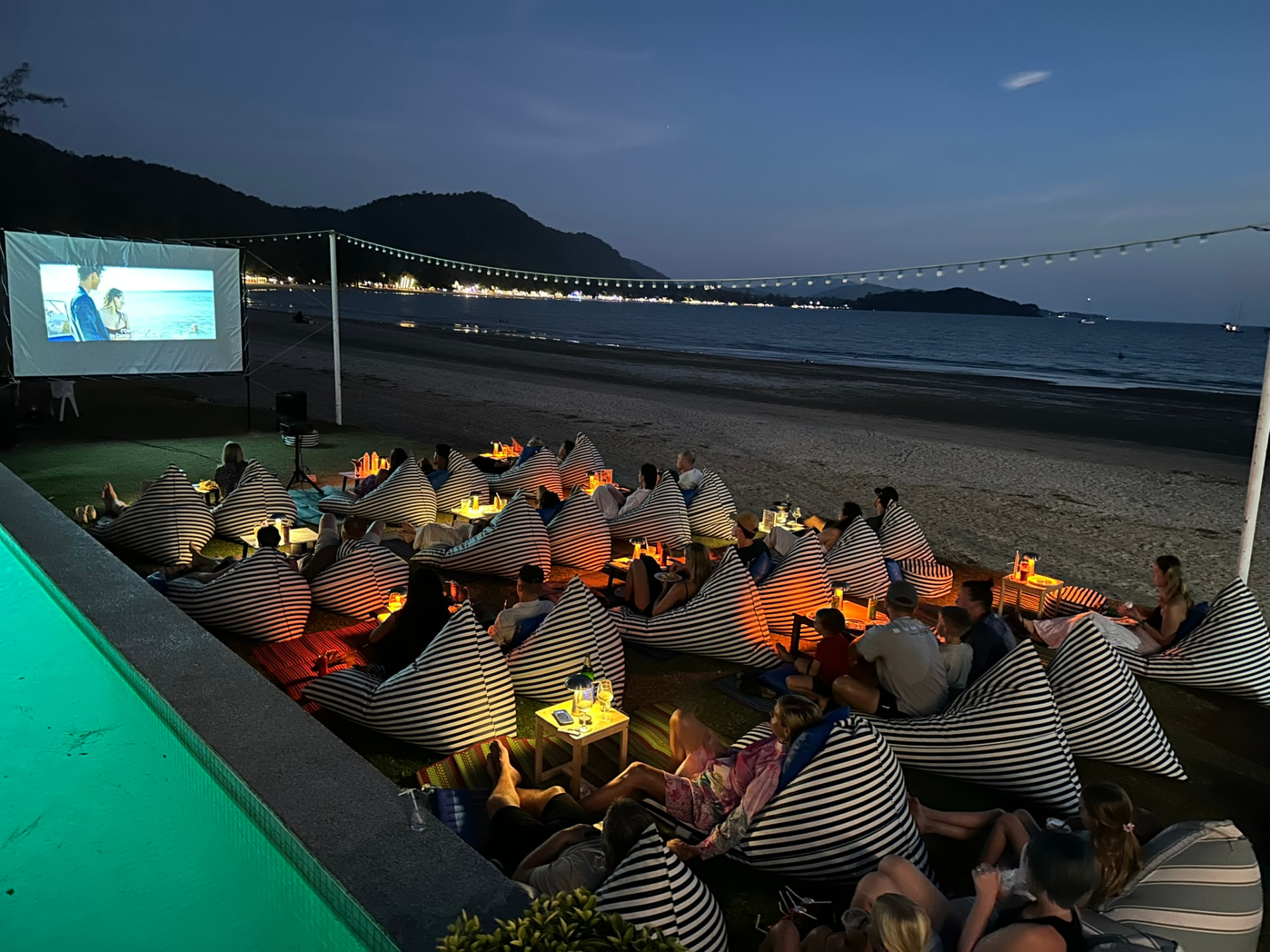 Guests watching a film on the beach during blue hour with lights across the bay
