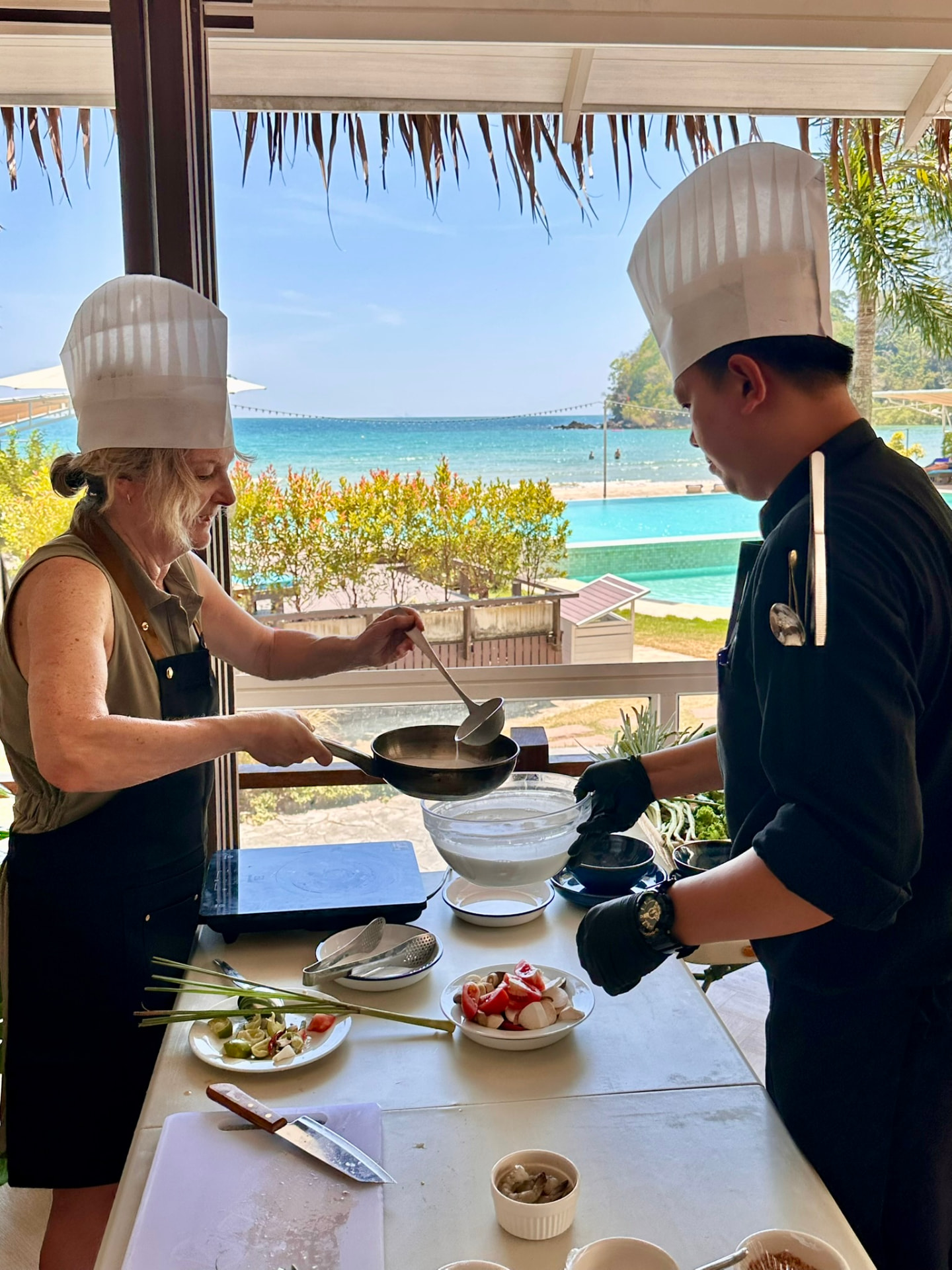 A guest cooking in a wok beside the chef with the beach behind