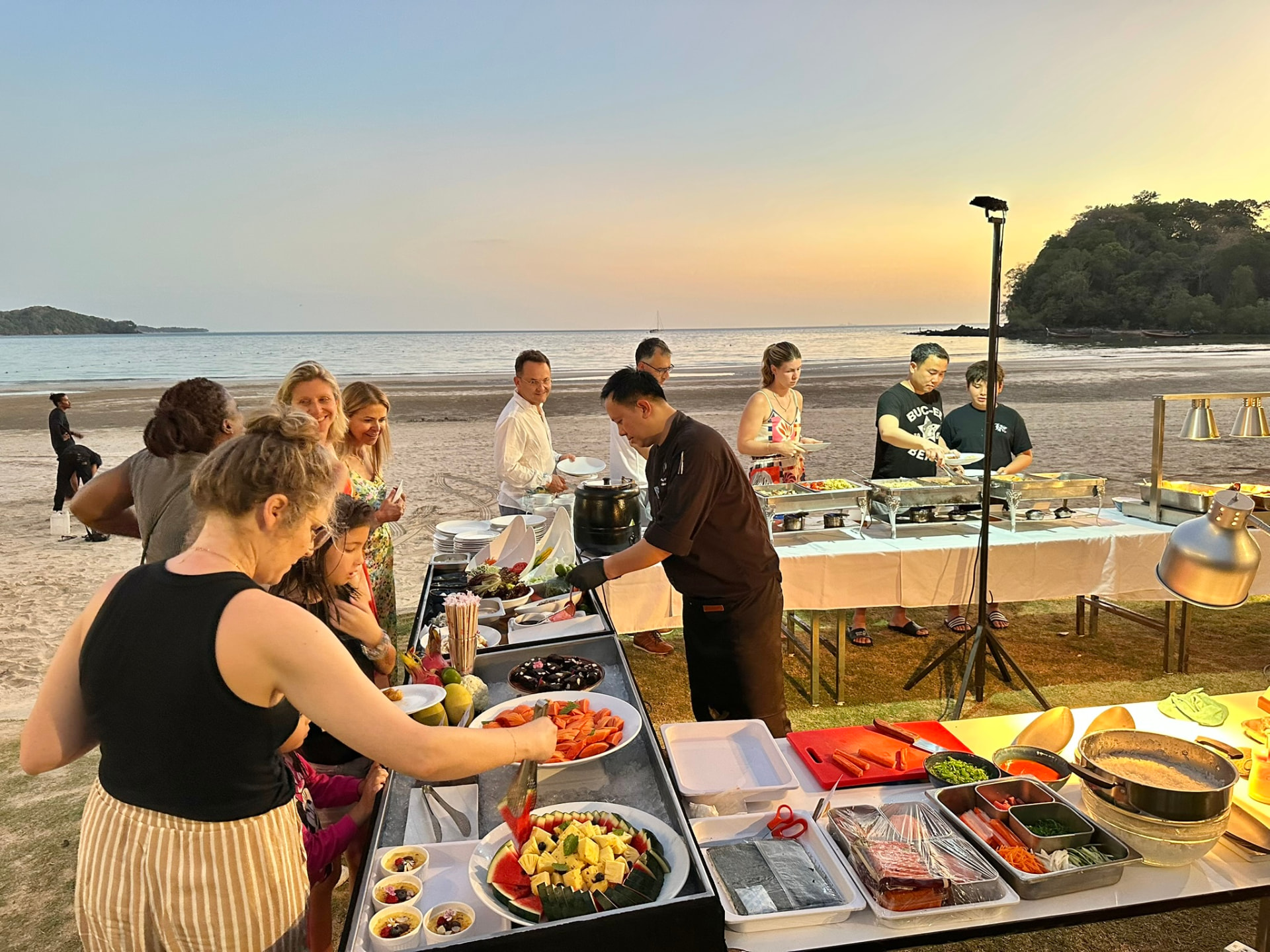 Guests and the chef at the beachside buffet with fresh fruit and seafood as the sun sets