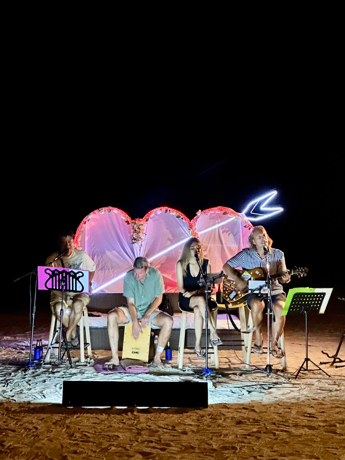 A four-piece band performing on the sand in front of a lit heart backdrop